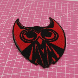 May include: A red and black owl-shaped embroidered patch. The patch features a stylized owl design with large, round eyes and a pointed beak. The owl's body is black, and the outline and details are red. The patch is likely for clothing or accessories.