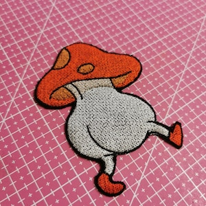 Könnte beinhalten: Gestickter Aufnäher mit einem skurrilen Pilzdesign. Der Pilz hat einen roten Hut mit orangefarbenen Punkten, einen weißen Körper und orangefarbene Stiefel. Der Aufnäher ist schwarz umrandet und eignet sich zum Aufbügeln oder Aufnähen auf Kleidung oder Accessoires.