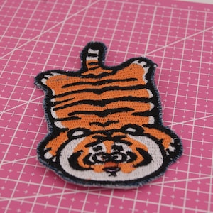 Pode incluir: Patch bordado com um desenho de tigre de desenho animado. O patch tem listras laranja e pretas, um rosto branco e um contorno preto. É exibido em uma esteira de corte rosa.