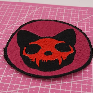 Könnte beinhalten: Gestickter Aufnäher mit einem schwarzen Katzenschädel-Design in Rot und Schwarz. Der Aufnäher ist kreisförmig mit einem dunkelrosa Hintergrund und einem schwarzen Rand. Der Katzenschädel hat scharfe Zähne.