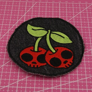 Op de afbeelding: Geborduurde patch met twee rode kersen met schedelgezichten, groene bladeren en stelen. De patch heeft een zwarte denim-achtige achtergrond en een gestikte rand. Een uniek ontwerp dat schattige en edgy elementen combineert.