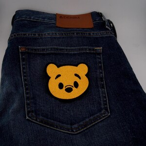Puede incluir: Vaqueros de mezclilla azul oscuro con un parche de oso amarillo en el bolsillo trasero. El parche presenta una nariz y ojos negros. Una etiqueta de cuero marrón con "& DENIM" es visible encima del bolsillo trasero.