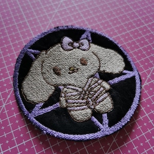 Può includere: Toppa ricamata raffigurante un coniglietto dei cartoni animati con un fiocco, che indossa un abito a righe, e inserita in un cerchio nero con un disegno a pentagramma viola. La toppa ha un bordo cucito ed è su un tappetino da taglio rosa.