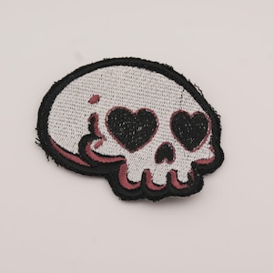 May include: Embroidered patch featuring a skull with heart-shaped eyes. The skull is white with black and burgundy accents, and a black border. This patch is suitable for jackets, bags, or other fabric items.