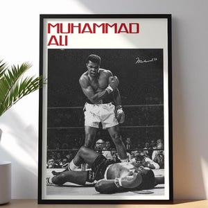 May include: Black and white framed print featuring Muhammad Ali standing over a defeated opponent in a boxing ring. The words "MUHAMMAD ALI" are printed in red at the top. A signature is visible on the right side.