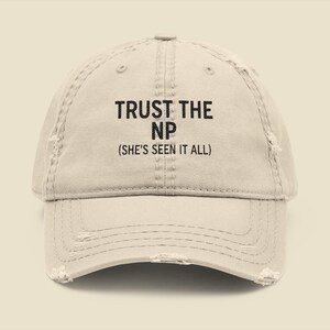May include: A distressed beige baseball cap with the text "TRUST THE NP (SHE'S SEEN IT ALL)" in black. The cap has a curved brim and a worn appearance, suggesting a vintage or well-loved style. The text is centered on the front.