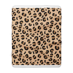 Könnte beinhalten: Eine rechteckige Wohndecke mit einem sich wiederholenden Leopardenmuster. Das Design besteht aus unregelmäßigen schwarzen und dunkelbraunen Flecken auf hellbraunem Grund, abgeschlossen mit einem weißen Rand an der Ober- und Unterseite.