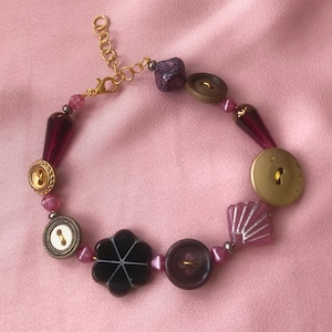 May include: A handmade bracelet featuring a variety of beads and buttons in shades of gold, brown, purple, and pink. The bracelet includes a gold-toned chain and clasp, with a mix of round, square, and flower-shaped elements.