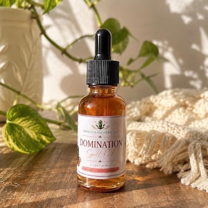 May include: An amber glass dropper bottle containing liquid, featuring a white label with the text Emerald Alchemy, LLC, Domination Spell Oil, and 1 oz / 30 ml. The bottle has a black dropper cap and is filled with a golden-colored oil.
