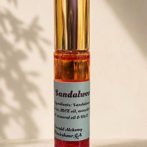 Minimalist sandalwood perfume oil roller bottle with a gold cap on a soft beige background, featuring warm natural lighting and delicate botanical shadows. Amber and red-toned oil bottle by Emerald Alchemy displayed in a clean aesthetic luxury product photography style.