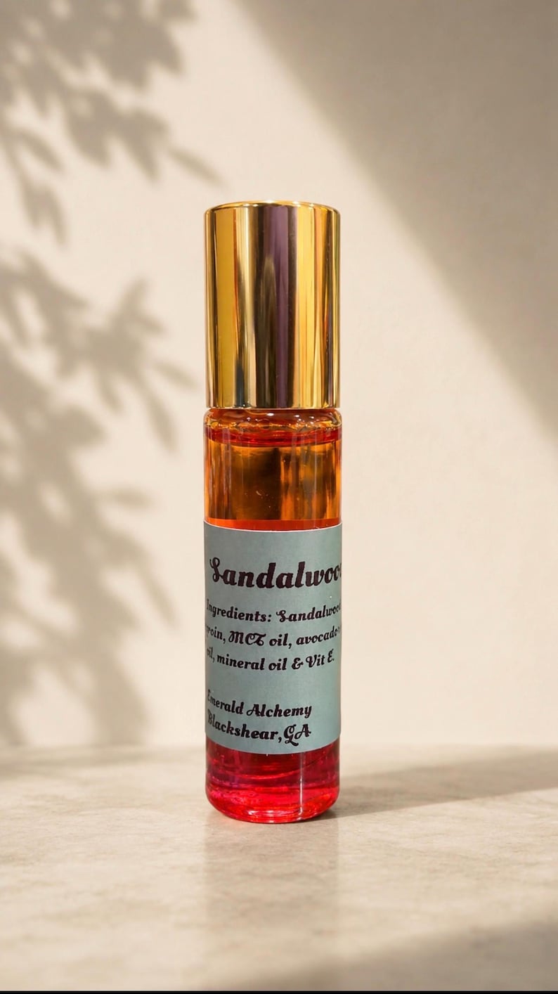 Minimalist sandalwood perfume oil roller bottle with a gold cap on a soft beige background, featuring warm natural lighting and delicate botanical shadows. Amber and red-toned oil bottle by Emerald Alchemy displayed in a clean aesthetic luxury product photography style.