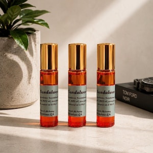 Three sandalwood perfume oil roller bottles with gold caps displayed on a bright neutral stone surface with soft natural sunlight, greenery, and modern masculine decor accents. Clean luxury product photography featuring Emerald Alchemy amber glass perfume rollers styled with warm lighting, earthy tones, and a sleek minimalist aesthetic.