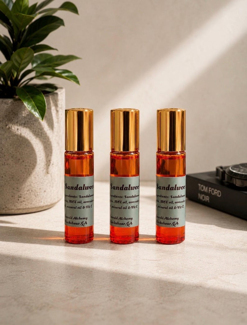 Three sandalwood perfume oil roller bottles with gold caps displayed on a bright neutral stone surface with soft natural sunlight, greenery, and modern masculine decor accents. Clean luxury product photography featuring Emerald Alchemy amber glass perfume rollers styled with warm lighting, earthy tones, and a sleek minimalist aesthetic.