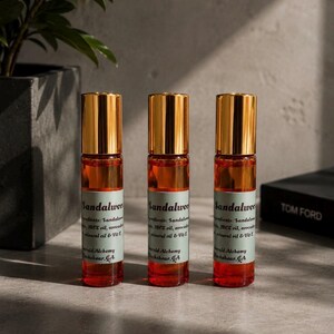 Three sandalwood perfume oil roller bottles with gold caps displayed on a modern stone surface with moody natural lighting, dark greenery, and minimalist masculine decor in the background. Clean luxury product photography featuring Emerald Alchemy amber glass perfume rollers styled in a sleek masculine aesthetic with warm shadows and earthy tones.