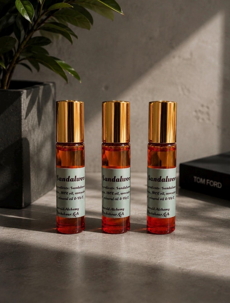 Three sandalwood perfume oil roller bottles with gold caps displayed on a modern stone surface with moody natural lighting, dark greenery, and minimalist masculine decor in the background. Clean luxury product photography featuring Emerald Alchemy amber glass perfume rollers styled in a sleek masculine aesthetic with warm shadows and earthy tones.