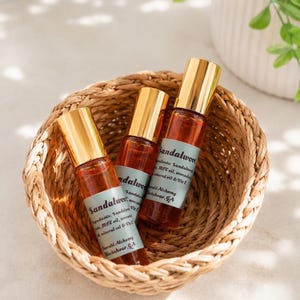 Three sandalwood perfume oil roller bottles with gold caps arranged inside a woven basket on a soft neutral surface, styled with bright natural lighting and subtle greenery in the background. Clean aesthetic flat lay product photography featuring Emerald Alchemy amber glass perfume rollers in a warm, minimalist spa-inspired setting.