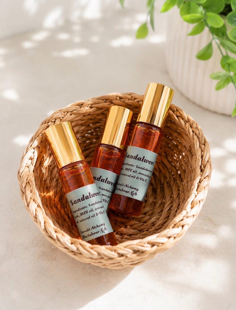 Three sandalwood perfume oil roller bottles with gold caps arranged inside a woven basket on a soft neutral surface, styled with bright natural lighting and subtle greenery in the background. Clean aesthetic flat lay product photography featuring Emerald Alchemy amber glass perfume rollers in a warm, minimalist spa-inspired setting.