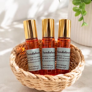 Three sandalwood perfume oil roller bottles with gold caps displayed in a woven basket on a bright neutral surface, surrounded by soft natural lighting and greenery accents. Clean aesthetic product photography featuring Emerald Alchemy amber glass perfume rollers in a warm, minimalist spa-inspired setting.