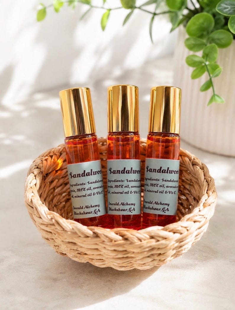 Three sandalwood perfume oil roller bottles with gold caps displayed in a woven basket on a bright neutral surface, surrounded by soft natural lighting and greenery accents. Clean aesthetic product photography featuring Emerald Alchemy amber glass perfume rollers in a warm, minimalist spa-inspired setting.