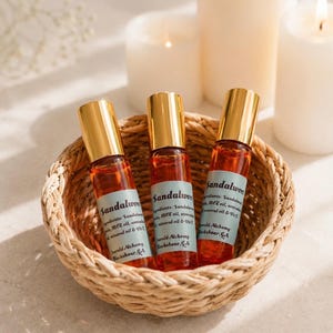 Three sandalwood perfume oil roller bottles with gold caps displayed in a woven basket beside glowing white pillar candles on a neutral stone surface. Warm natural lighting and soft shadows create a clean luxury spa aesthetic with Emerald Alchemy amber glass perfume rollers in a calming minimalist setting.