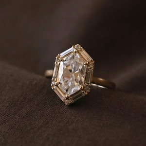 May include: A gold ring featuring a large, elongated hexagonal center stone surrounded by a halo of smaller, rectangular baguette-cut stones and tiny round accent stones, set on a thin, polished gold band.
