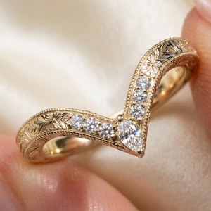 May include: A V-shaped gold ring featuring a central marquise-cut diamond flanked by round brilliant-cut diamonds. The band is crafted with intricate floral engraving and milgrain detailing along the edges, creating a textured finish.