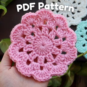 May include: A close-up of a pink, crocheted coaster with a floral design. The coaster is round with a scalloped edge and openwork details. The text "PDF Pattern" is in white at the top.