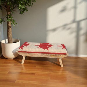 Peut inclure: Banc en bois avec une assise rectangulaire ornée d'un motif de feuille d'érable rouge sur fond crème. Le banc a quatre pieds inclinés et une bordure rouge. Le banc mesure environ 91 cm de long.