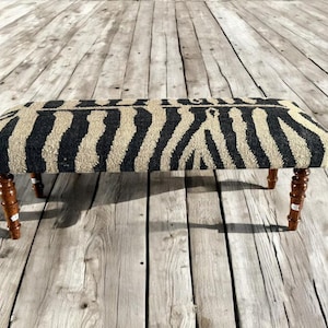 Puede incluir: Un banco rectangular con un estampado de cebra en negro y beige. El banco tiene cuatro patas de madera torneada de color marrón oscuro. El banco está colocado sobre un suelo de madera.