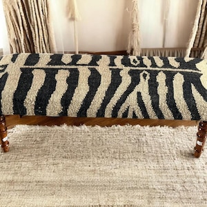 Könnte beinhalten: Eine rechteckige Bank mit einem Zebrastreifenmuster in Schwarz und Beige. Die Bank hat vier gedrechselte Holzbeine. Die Bank steht auf einem beigen Teppich.