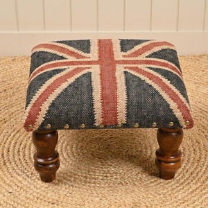 May include: A small, square footstool featuring the Union Jack flag design. The top is upholstered in a textured fabric with the red and white stripes and cross over a dark gray background. The wooden legs are dark brown.