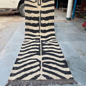 May include: A rectangular rug with a zebra stripe pattern in black and beige. The rug has a fringe on the bottom edge and a design resembling a tiger's face at the top. The rug is made of a woven material.