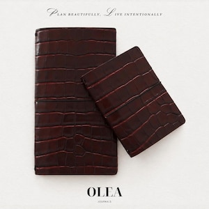 May include: Two dark burgundy journals with a glossy crocodile-embossed texture, each secured with a thin black elastic band. The text at the top reads "PLAN BEAUTIFULLY, LIVE INTENTIONALLY" and the text at the bottom reads "OLEA JOURNALS."