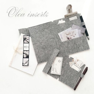 Peut inclure: Organisateur en feutre gris avec plusieurs poches, une pince métallique et un porte-stylo. L'organisateur contient diverses cartes et papiers. Le texte "Olea inserts" est affiché en haut à gauche. Un bloc-notes blanc avec un ruban est à gauche.