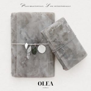 May include: Two gray marble-patterned journals with elastic bands. The larger journal features four charms: a white shell, a clear crystal, a dark green stone, and a white scallop shell. Text reads: PLAN BEAUTIFULLY, LIVE INTENTIONALLY and OLEA JOURNALS.