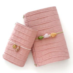 May include: Two pink, textured leather journals. The smaller journal has a white cord with two gold charms and a pink crystal pendant. The larger journal is secured with a green cord adorned with colorful beads. The journals are set against a white background.