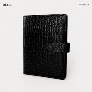 May include: A black journal with a glossy crocodile-embossed texture and a matching side strap closure. The cover features a repeating scale pattern. The text OLEA JOURNALS is printed in the top corners of the frame in a clean, black font.