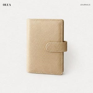 May include: A beige journal with a textured lizard-skin pattern and a matching side strap closure. The cover features a vertical orientation with stitched edges. The text OLEA is printed in the top left corner and JOURNALS in the top right.