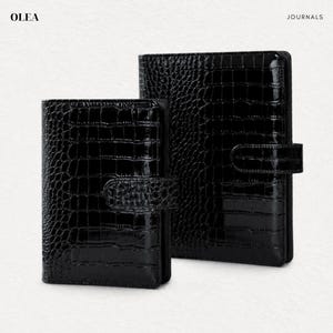 May include: Two black journals with a glossy crocodile skin texture and a secure side strap closure. The text OLEA JOURNALS is printed in the top corners. These rectangular notebooks feature a dark, reflective finish and a structured design.