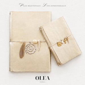 May include: Two gold-colored journals with metallic covers. The larger journal features a silver elastic band with a stone slice, a silver spiral charm, and two beads. The smaller journal has a silver band with a star charm, a pearl, a cross, and a tooth-shaped charm. Text reads: PLAN BEAUTIFULLY, LIVE INTENTIONALLY and OLEA JOURNALS.
