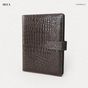 May include: A dark brown journal with a glossy crocodile skin texture and a matching side strap closure. The cover features a repeating embossed scale pattern. The text OLEA JOURNALS is printed in the top corners of the white background.