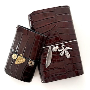 May include: Three dark brown leather journals with a crocodile skin texture. The journals are decorated with charms, including a gold heart, a sunburst, and a silver flower. The journals are tied with black elastic.