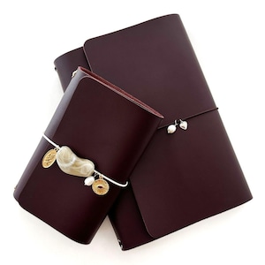 May include: Burgundy leather journals with a wrap closure. One journal has a white cord with a stone, gold coin, and pearl charm. The other journal has a silver elastic closure with pearl and heart charms. The journals are on a white background.