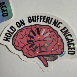 May include: A holographic sticker with the words "HOLD ON BUFFERING ENGAGED" surrounding a pink brain illustration. The brain graphic has a loading symbol. The sticker has a white border and a shiny, iridescent finish.