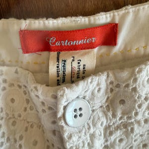 May include: Close-up of white eyelet shorts with a button closure. The shorts have a decorative floral pattern and a red tag that reads "Cartonnier". A second tag is also visible.