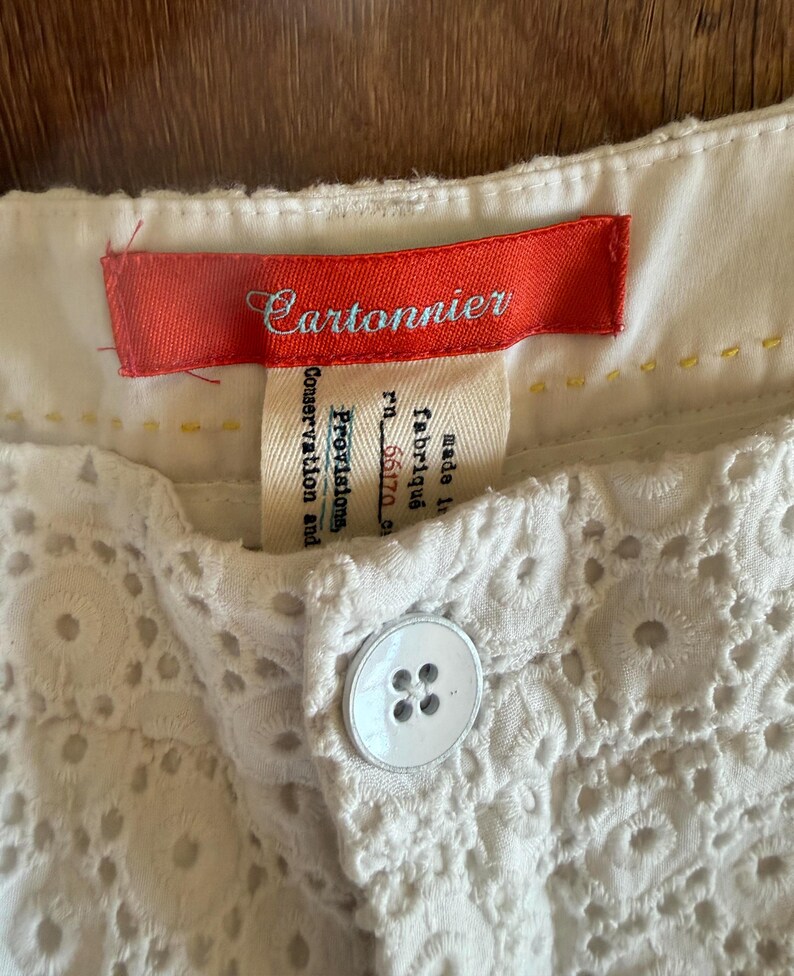 May include: Close-up of white eyelet shorts with a button closure. The shorts have a decorative floral pattern and a red tag that reads "Cartonnier". A second tag is also visible.