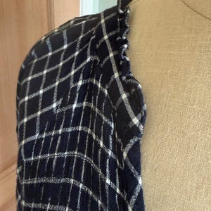 May include: A navy blue and white plaid patterned shirt sleeve with a ruffled edge. The fabric is draped over a mannequin, showcasing the texture and design of the garment. The plaid pattern is a classic design.