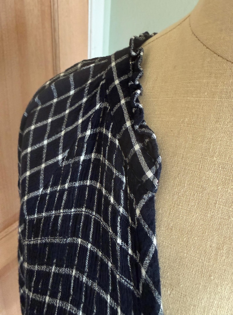 May include: A navy blue and white plaid patterned shirt sleeve with a ruffled edge. The fabric is draped over a mannequin, showcasing the texture and design of the garment. The plaid pattern is a classic design.