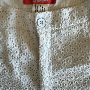 May include: Close-up of white eyelet shorts with a button closure. The shorts feature a detailed circular pattern throughout the fabric. A red tag with the brand name "Cartonnier" is visible at the waistband.