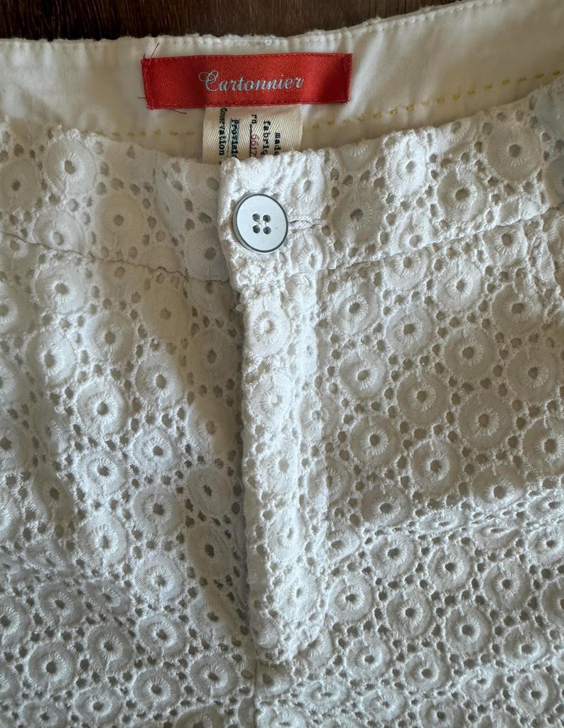 May include: Close-up of white eyelet shorts with a button closure. The shorts feature a detailed circular pattern throughout the fabric. A red tag with the brand name "Cartonnier" is visible at the waistband.